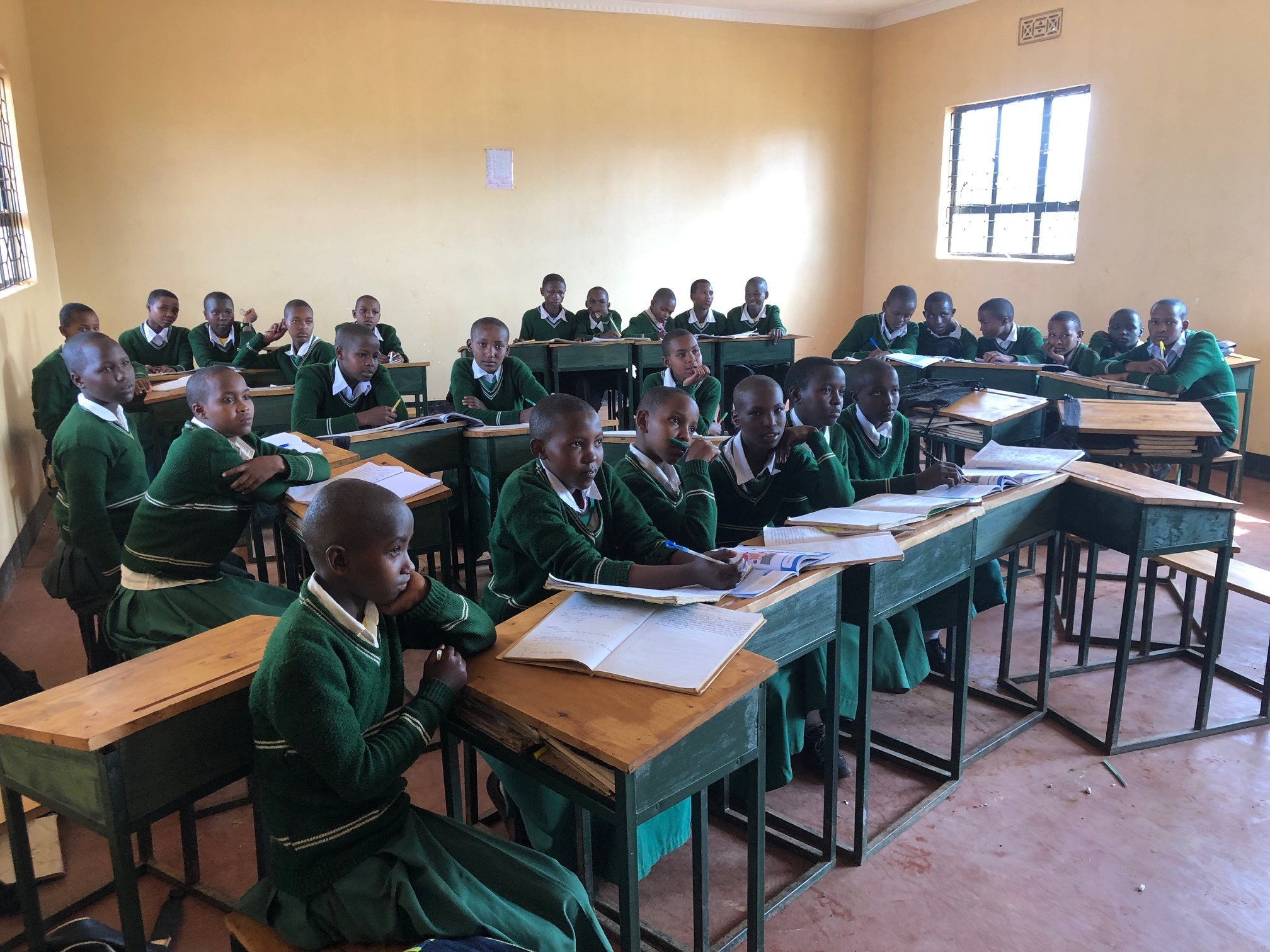 A classroom with students in green uniforms sit attentively at wooden desks arranged in a U-shape. Sunlight streams through a window, creating a bright, focused atmosphere.