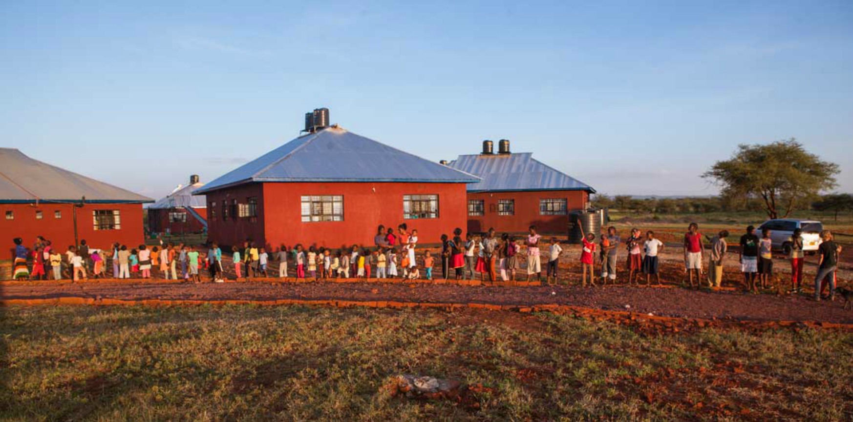 Children line up outside modern, red-roofed buildings in a rural setting, waiting for access to resources or services.