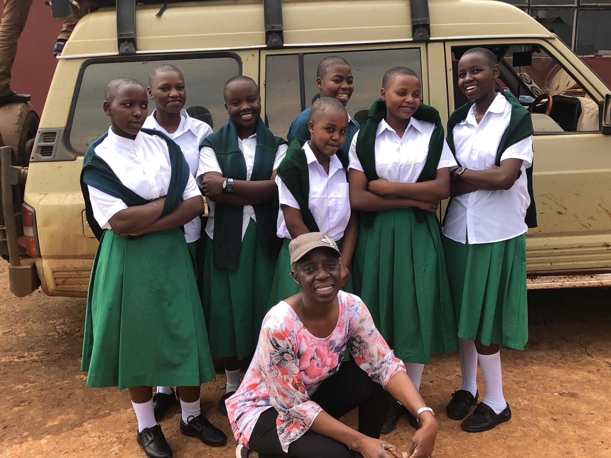 Seven smiling girls in white shirts and green skirts stand with arms crossed, posing in front of a dusty vehicle. A woman in a floral shirt and cap kneels in front, smiling warmly. The setting feels positive and joyful.