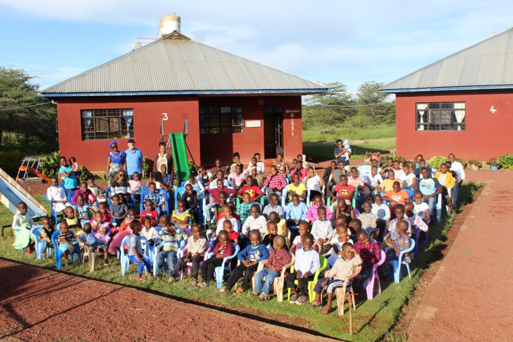 A large group of children sit on colorful plastic chairs outside red buildings, enjoying a sunny day, likely gathered for an organized event or celebration.