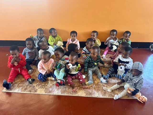 A group of toddlers sitting on a rug in two rows against an orange wall. They are dressed in colorful outfits, holding stuffed animals and toys, looking content.