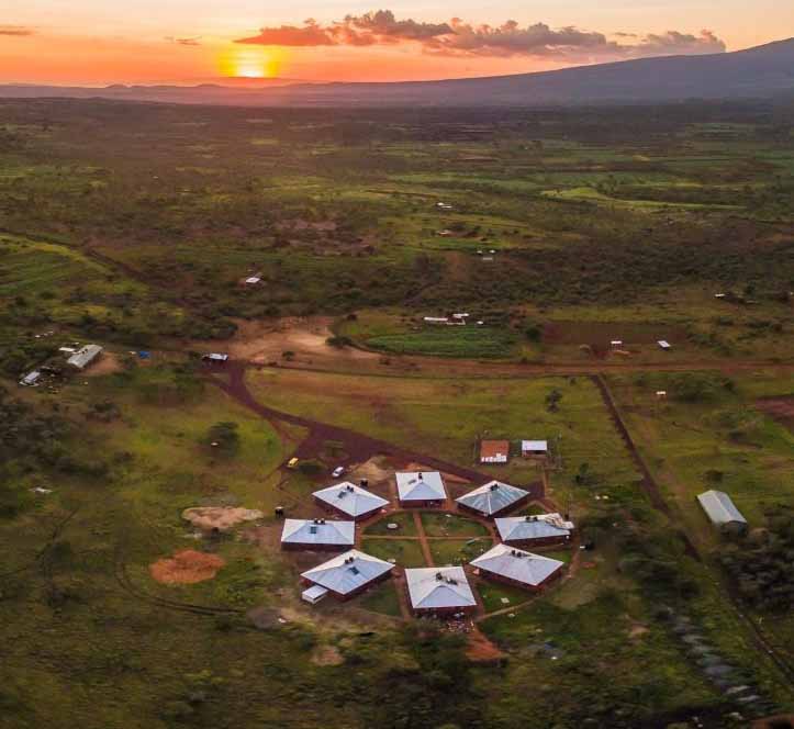 Aerial view of a circular village with hexagonal huts in a grassy landscape at sunset. The sky is orange, casting a warm glow over the scene.