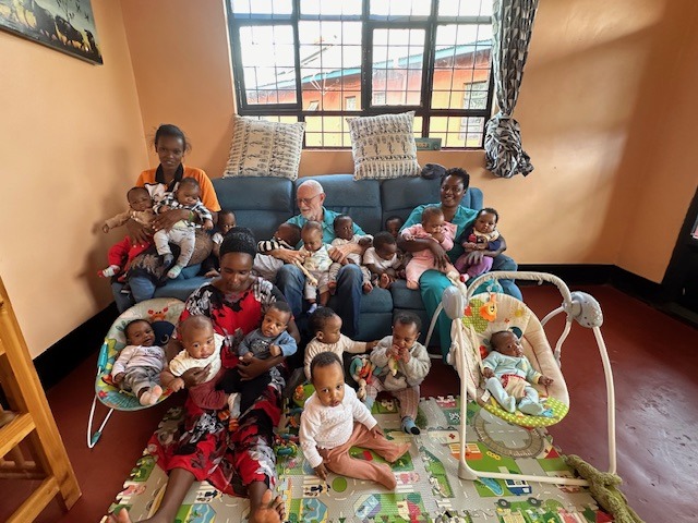 A cozy living room with three caregivers and numerous babies. Some babies are held, others are sitting or reclining. The scene conveys warmth and nurturing.